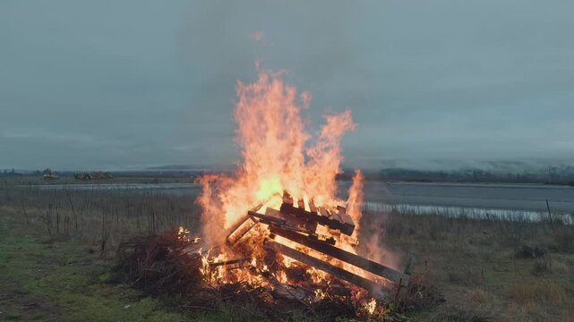 A controlled burn consumes pruned branches and wooden debris in a vineyard at dusk, with flames rising against a calm rural landscape during seasonal farm maintenance.
