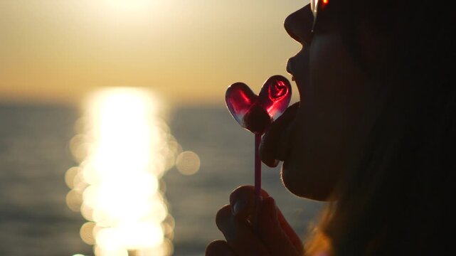 Heart-shaped candy sweetens a seaside sunset as a young woman enjoys a sugary treat. Static side profile close-up shot.