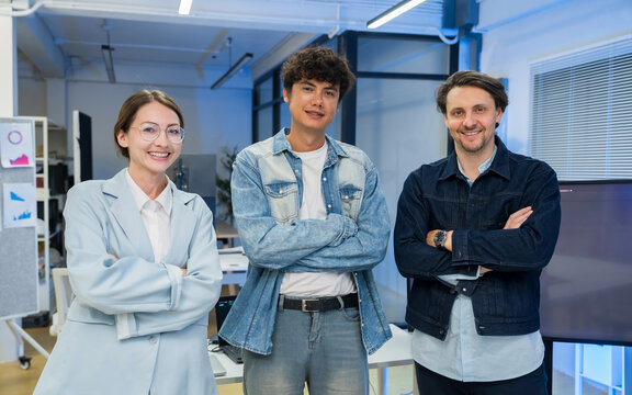 Portrait of a smiling diverse team of three business professionals standing with arms crossed in a modern office. They project confidence, teamwork, and unity in a smart casual work environment.