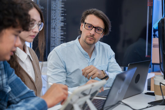 Team of three software developers discusses AI agent software while reviewing data and interface graphics on laptops in modern technology office, collaborating develop intelligent digital solutions.