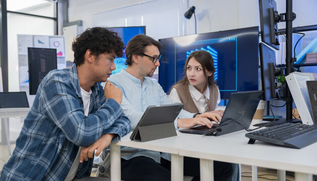 Team of three software developers discusses AI agent software while reviewing data and interface graphics on laptops in modern technology office, collaborating develop intelligent digital solutions.