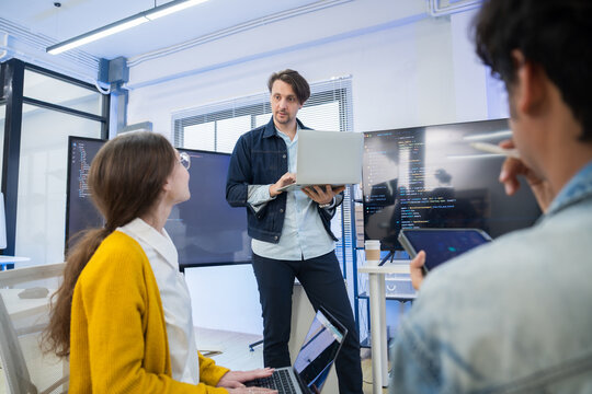 Caucasian businessman holding laptop and presenting software updates to seated team members in office.