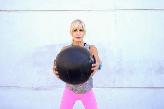 Fit woman holding medicine ball at chest, standing ready for strength exercise against pale wall with calm daylight and focused posture before squat or throw movement