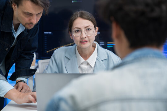 Focused Caucasian female engineer in glasses speaking with multiracial team members around laptop desk in office.