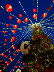 Christmas tree decorated with numerous red lanterns and sparkling lights, prominently displaying a panda ornament, all set against a deep blue evening sky creating a festive atmosphere © Irina Mikhailichenko