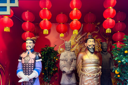 Traditional Chinese warrior statues in vibrant colors stand prominently against a red backdrop adorned with hanging lanterns and decorative plants, creating a festive atmosphere for Lunar New Year