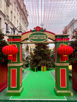 Festive archway adorned with red lanterns and a sign for Chinese New Year, featuring a decorated Christmas tree and colorful gifts, set against an urban backdrop with a green carpet