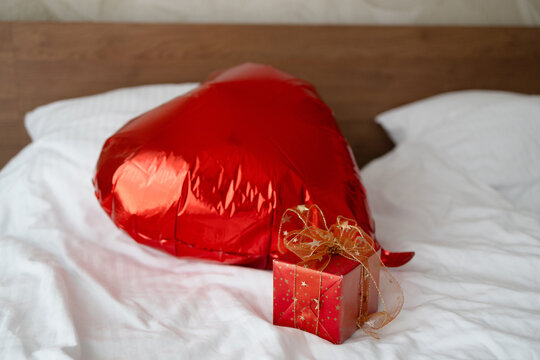 Close up of a red foil heart balloon and small wrapped present with gold ribbon on white sheets in bedroom. Concept of Valentine's Day surprise, romantic holiday celebration, and honeymoon hotel room