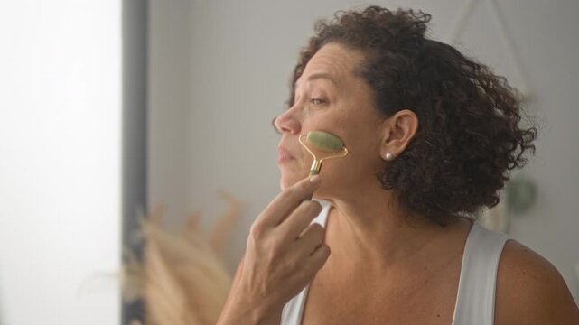 Woman rolling a jade face roller along her chin and cheek while watching her reflection in a mirror inside a building; self care calm wellness.