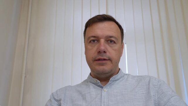Mans neutral expression in office setting, observing viewer directly against subtly lit drape curtain, hinting at internal monologue or moment of contemplation.