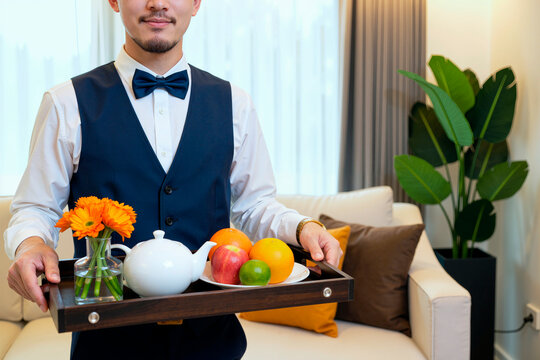 Portrait of Asian young adult waiter holding serving tray with tea set, fruit, flowers in hotel room, presenting hospitality service. Ideal for hotel marketing, room service, travel ads
