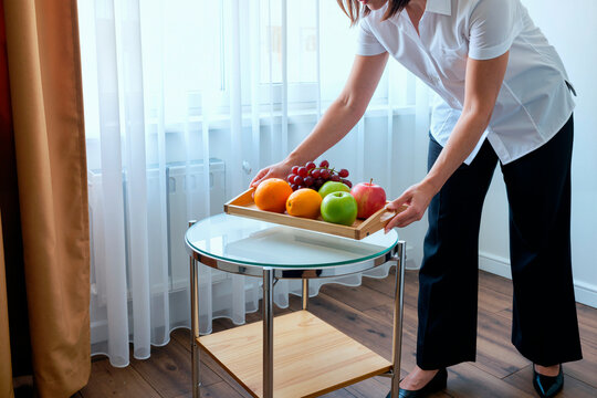 Caucasian young adult woman placing tray with fresh fruit on table in hotel room, preparing healthy welcome service. Useful for hospitality, wellness, nutrition marketing campaigns