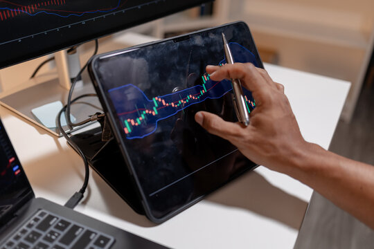 Professional trader analyzing stock market charts across multiple screens using laptop and tablet to monitor price movements, technical indicators and investment strategies in modern trading workspace