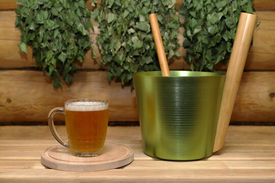 On a wooden bench, against the backdrop of a log wall with dry birch brooms hanging on it, there are a mug of light beer on a round wooden tray and a metal bucket with a ladle.