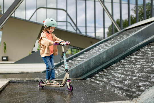 Happy young boy with a kick scooter playing beside a modern urban fountain. Active child wearing a safety helmet enjoys outdoor recreation and water in the city.