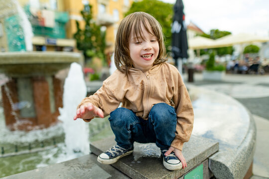 Playful young boy having fun beside public fountain in the city. Happy child reaches toward splashing water and laughs during a carefree outdoor summer day.