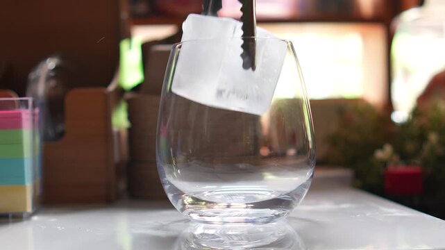 4K Close-up tongs dropping large square ice cube into clear glass on table in cafe, beverage preparation cocktail making refreshing drink and bar menu.