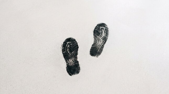 Two black shoe prints rest on pale surface, detailed tread patterns emphasize human presence, contrast highlights pause, stillness, and evocative trace of movement.