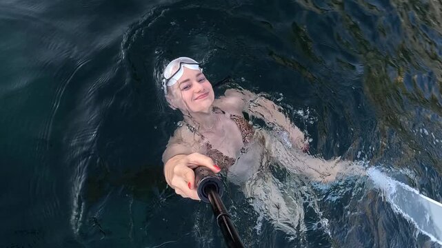 Self-recording woman floating in ocean water while holding monopod selfie stick. High-angle tracking shot.