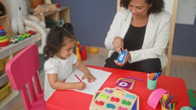 Teacher woman holds letter flashcard and writes with pen while little girl draws with pencil at preschool classroom; literacy nurturing focus.