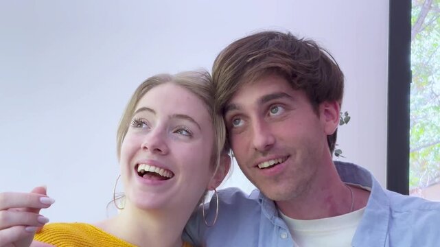 A happy young couple points and looks with excited expressions from their sofa. Sharing a joyful, heartwarming moment, they laugh together, embodying affection and connection in their home.