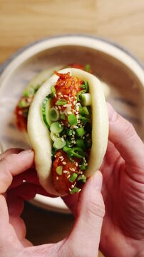 Hands hold hot shrimp bao with spicy glazed shrimp. Shrimp bao is topped with scallion and sesame seeds. Enjoy shrimp bao as quick seafood snack with chilli dip, cucumber salad, noodles or rice.