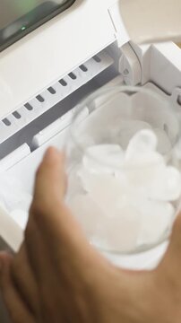 Vertical video of hand taking ice cubes from ice maker machine and pouring into glass