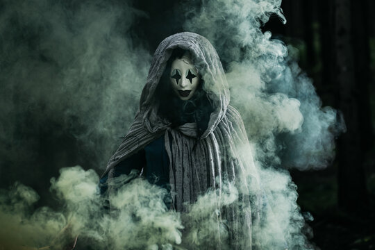 Spooky and scary Halloween themed image in a dark forest with smoke and an eerie woman with a mask. .
