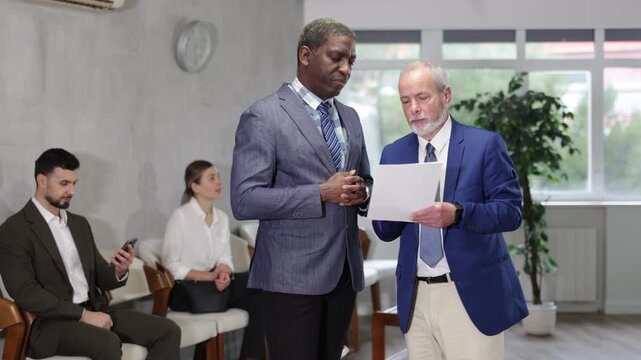 Eldery businessman holds documents and talks to adult man job seeker in office lobby