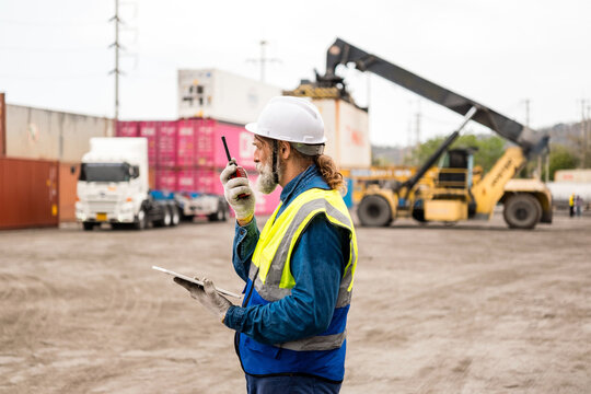 Safety-vested worker with hard hat uses a radio and tablet at a container yard while a reach stacker and truck move shipping containers in the background