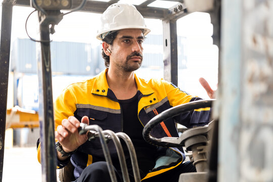 Warehouse worker driving a forklift, wearing a hard hat and high-visibility jacket, focused on the controls amid shipping containers in an industrial yard.