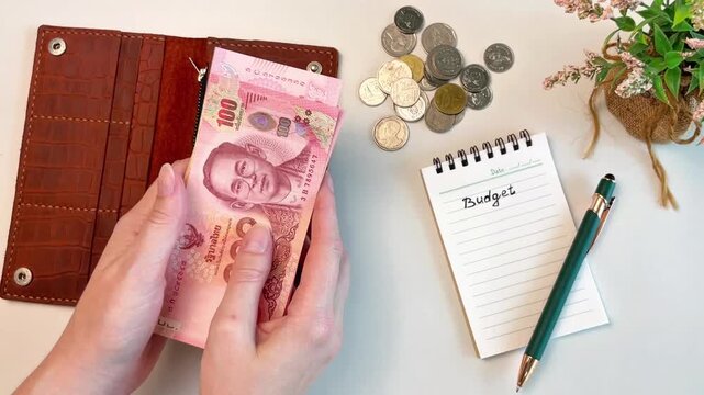 A close-up of a woman's hands counting Thai baht banknotes next to a wallet, coins, and a notepad labeled "Budget." Conceptual concepts of personal finance, income, expenses, and budgeting.