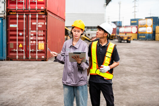 Two workers in helmets and safety vests review shipping plans on a tablet at a container yard, with stacked cargo containers and trucks in the background.