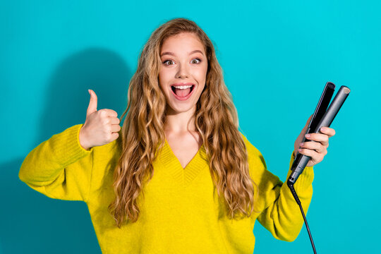 Young woman in yellow sweater giving thumbs up holds hair straightener in bright blue studio background