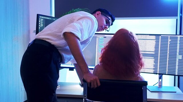 Two colleagues collaborate at a workstation with multiple monitors showing code, as a mentor leans in to guide a developer in a blue-lit office.