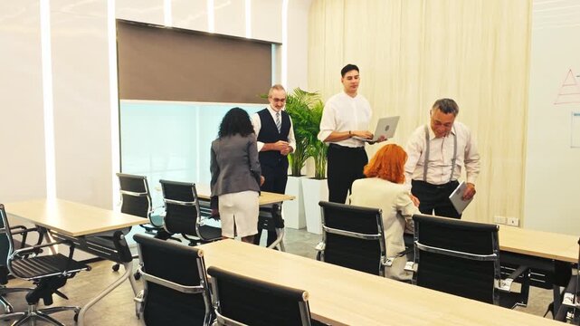 A group of coworkers gathers in a modern conference classroom, chatting before a training session amid rows of desks and rolling chairs, with a whiteboard at the front