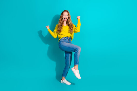 Young woman jumping with joy in a bright yellow sweater jeans and white sneakers against a blue background