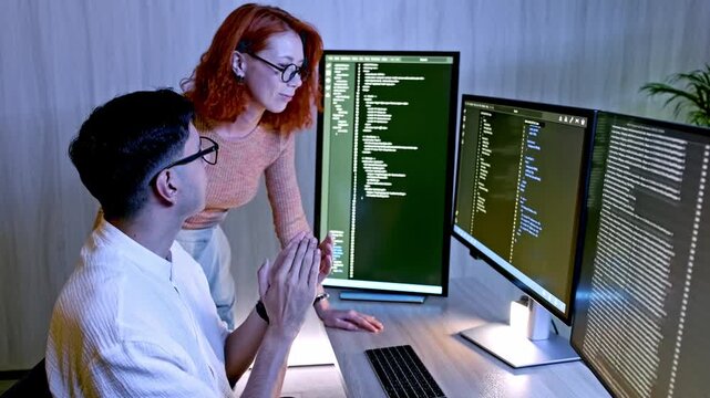 Two developers collaborate at a desk with multiple monitors, reviewing and debugging code in a modern office.