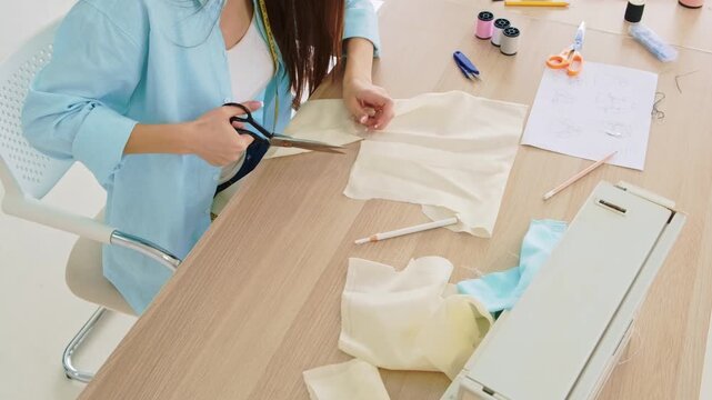 Person cutting fabric on a worktable beside a sewing machine, thread spools, and pattern sketches in a bright studio, preparing to sew a garment.