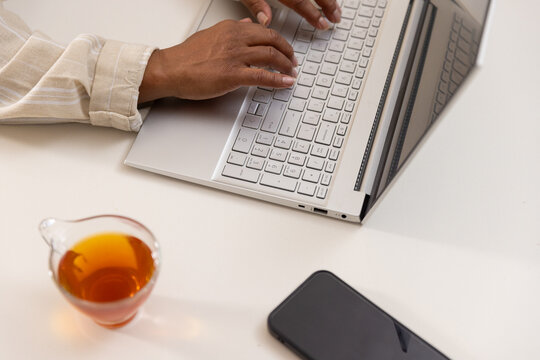 African American woman typing on silver laptop at home desk with phone, tea, cuffed sleeve