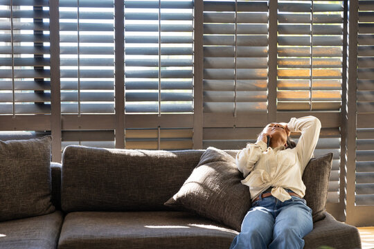 African American woman sitting on sofa tied shirt jeans pillow and shutters sunlight, copy space