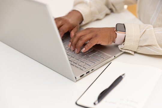 Mature African American woman typing on silver laptop at desk wearing smartwatch, notepad and pen