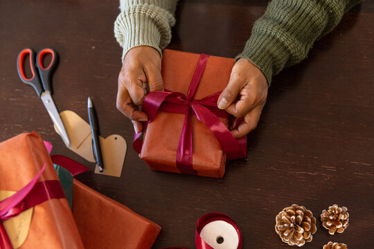 Senior African American woman in knit sleeves tying burgundy ribbon on wrapped gift at home table