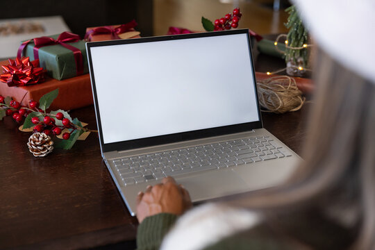 African American woman in green sweater, red hat typing at wood table with laptop, gifts, twine