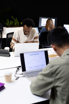 Diverse coworkers analyzing laptop charts at office desk with tablet, man wearing cream sweater