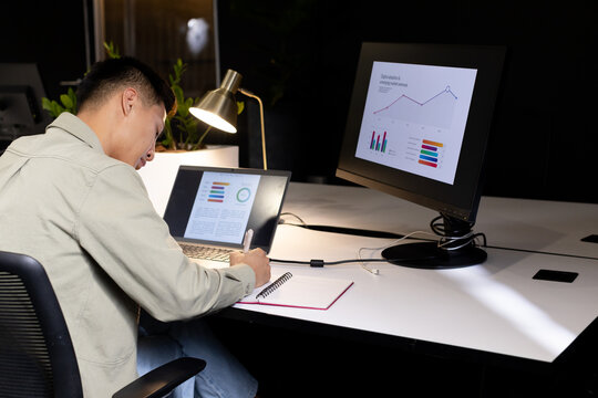 Asian man wearing khaki jacket writing in spiral notebook at office with laptop, monitor