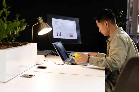 Mid-adult Asian man sitting, working on laptop and monitor in office holding green mug, copy space