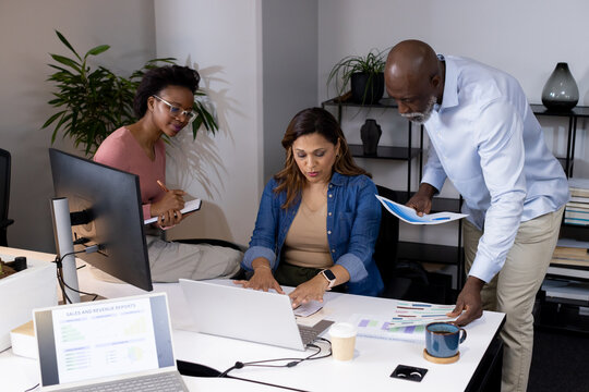 Office team collaborating around white desk at modern office reviewing laptop and printed charts