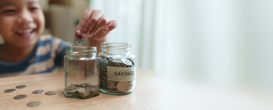 Happy child placing a coin into glass savings jars at home with copy space, representing money saving, budgeting, financial planning, savings goals and responsible financial habits.