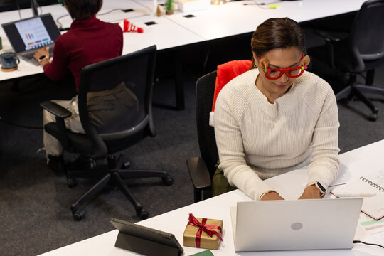 Female coworkers wearing cream sweater, red glasses typing on laptop at office with wrapped gift